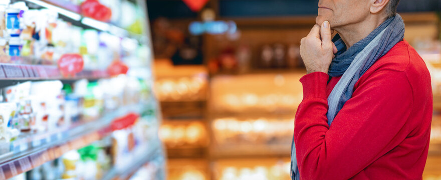 A man stands in a grocery store looking at shelves filled with food items. He appears to be lost in thought as he decides what to buy. The store is well-lit and busy with other shoppers.