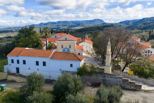 Aerial view of Pantokrator Monastery in Agios Athanasios, Corfu, Greece. Beautiful yellow church, traditional white buildings with red roofs, and scenic green mountains under a cloudy blue sky.