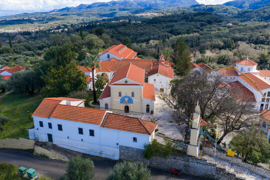 Drone shot of the historic Pantokrator Monastery complex in Agios Athanasios, Corfu, Greece. Capturing the central courtyard, detailed church mosaic, bell tower, and lush Mediterranean nature.