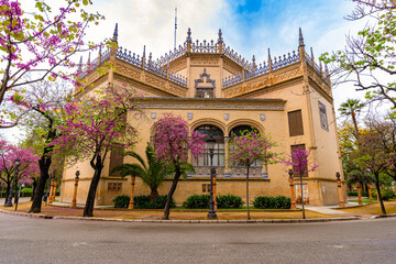 Naklejka premium Front view of a historic Neo-Mudejar building in Seville with intricate brickwork, arched windows, and decorative crest under a vibrant cloudy sky.