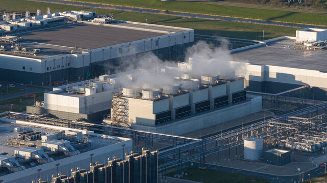 Aerial view of the cooling towers of a large industrial data center facility with steam rising against a backdrop of modern buildings and green fields in Eemshaven, Groningen, Netherlands.