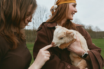 Obraz premium Lifestyle scene of two women outdoors with a gentle goat kid, showcasing affectionate interaction, joyful bond, warm light on a rural meadow, candid moment of care and friendship in natural colors,