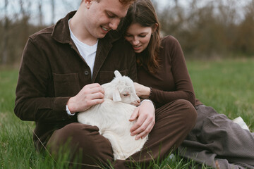 Obraz premium Couple sits on grass in a meadow with a gentle goat kid resting on their laps, warm affectionate moment outdoors, casual rustic clothing, calm atmosphere, natural light, true lifestyle scene with