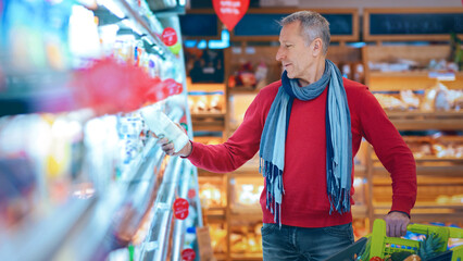 A man examines a bottle while grocery shopping. He stands in the aisle of a store with shelves filled with products. A shopping cart is beside him. © Prostock-studio