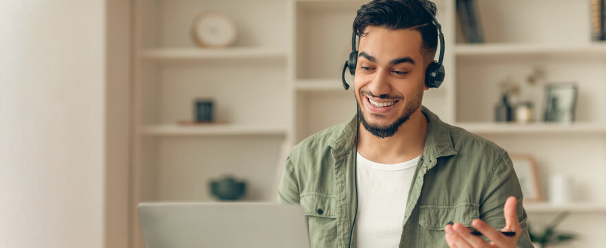 A man is sitting at a table in a modern room. He is using a laptop and wearing a headset. He appears to be talking and smiling, engaged in an online meeting.