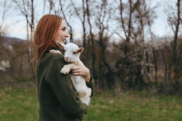 Obraz premium A woman in a green sweater gently cradles a young goat in a sunlit field, smiling. A candid moment of animal friendship, rural lifestyle, and calm countryside living.