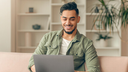 A young man sits on a couch with a laptop in front of him. He smiles as he works. The living room has bookshelves and plants around him. The setting looks bright and inviting. © Prostock-studio
