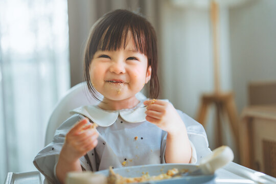 Happy cute Asian toddler girl eating and laughing in a high chair with messy face at home