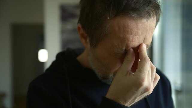 Middle-aged man with headache and painful grimace reacting to daylight at window, migraine symptoms and light sensitivity at home, 4K