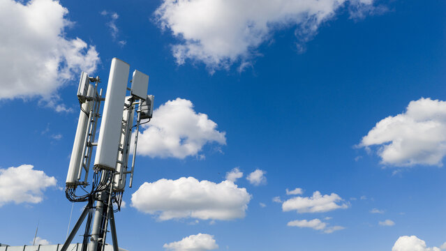 Aerial view of a telecommunications antenna array and cellular base station equipment against a bright blue sky with white clouds in Amsterdam, North Holland, Netherlands.