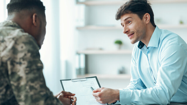 Two men are seated in an office setting. One man in military attire talks to another man in a blue shirt. They are engaged in conversation about a document.