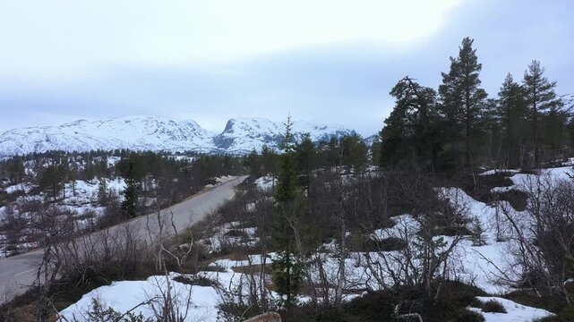 Forward drone movement above remote mountain road crossing snow-covered plateau at Hallbjonnsekken, Telemark during seasonal transition in Norway