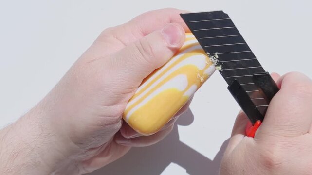 a hand cuts a yellow bar of soap with a utility knife on a white background, chunks and crumbs fly away as the blade cuts the soft soap, satisfying ASMR close-up.