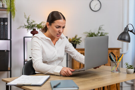 Young businesswoman at home office sees laptop freeze presses keys and shakes head in despair worry. Freelancer girl at table tries force restart unplugs cable waits and drafts support note for tech.