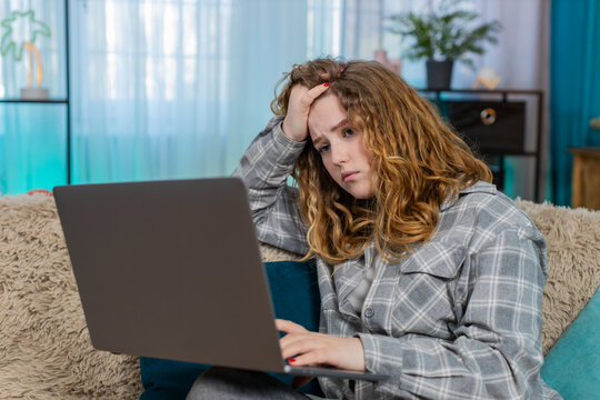 Young woman at home sitting with laptop feeling disappointed holding hand on forehead after failed attempt. Caucasian redhead girl on sofa frustrated, emotional reaction to unresolved online problem
