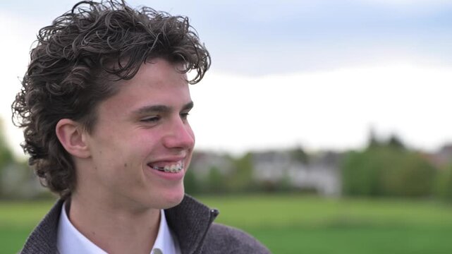 Close-up of smiling young man with braces talking outdoors in soft natural light. Happy male looking away, showing healthy teeth and positive эмоtions in nature.

