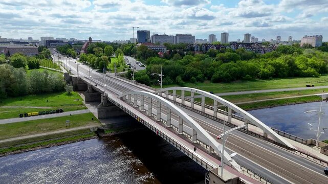 Aerial panorama of Poznan featuring Saint Roch Bridge over Warta River, white arched steel construction with tram tracks, green riverbanks, flowing water, urban cityscape background under sunny sky wi