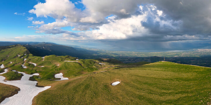 Aerial view of Le Colomby de Gex with green mountain ridges, patches of snow, and dramatic rain clouds over the valley near Echenevex, Auvergne-Rhone-Alpes, France.