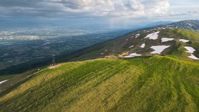 Aerial view of Le Colomby de Gex summit with green mountain ridges, melting snow patches, and the valley below under a dramatic cloudy sky Echenevex, Auvergne-Rhone-Alpes, France.