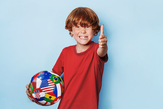 Red-haired child with soccer ball covered with multiple national flags smiling and shows likes with his fingers in the camera isolated blue background.