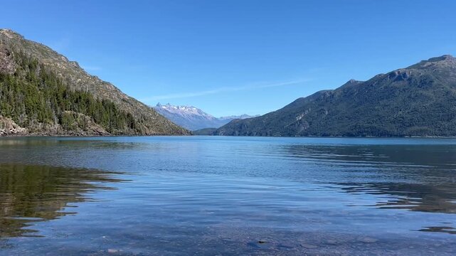 Crystal-clear blue water of Lago Puelo National Park filmed from a low angle almost at the water surface, creating an immersive perspective across the calm lake. The scenic landscape of Lago Puelo.