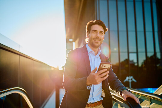 Young businessman smiling. Looking ahead while holding a smartphone and riding an escalator outdoors. Navigating the modern city at sunrise. Representing professional communication and urban lifestyle