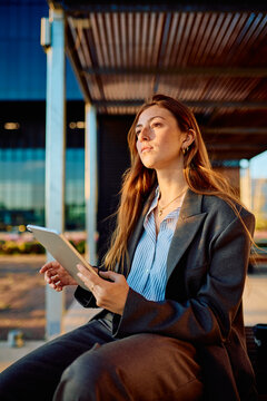 Young business professional woman holding a digital tablet and looking with a visionary expression in an urban setting during golden hour, embodying success and career growth
