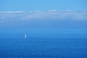 Fototapeta premium Minimal seascape with a sailboat and motorboat crossing calm blue ocean under soft clouds on Tenerife. Marine travel background with vast copy space