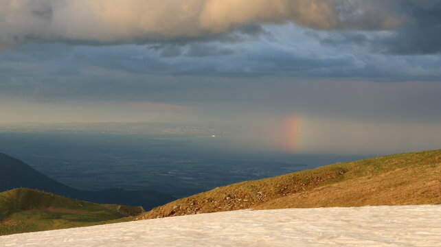 Aerial view of Le Colomby de Gex mountain ridge with snow patches, a rainbow over the valley, and dramatic storm clouds at sunset near Echenevex, Auvergne-Rhone-Alpes, France.