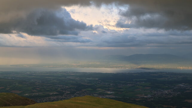 Aerial view of Le Colomby de Gex overlooking the valley and Lake Geneva under dramatic storm clouds with light rays in Echenevex, Auvergne-Rhone-Alpes, France.