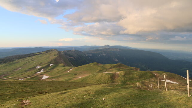 Aerial view of Le Colomby de Gex mountain ridge with green grassy slopes, patches of melting snow, and a wooden fence under a cloudy sky Echenevex, Auvergne-Rhone-Alpes, France.