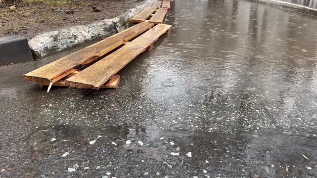 Wooden planks lie wet, slumped on icy, rain-slicked pavement. Rain falls steadily, pooling on cracked asphalt. Frost melts slowly beneath the wet surface. No people, no animals visible
