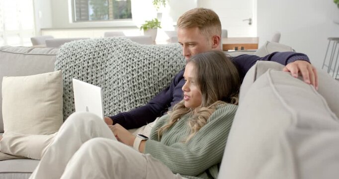 Diverse couple sitting on sofa white laptop drawing woman into typing and scrolling as man watching