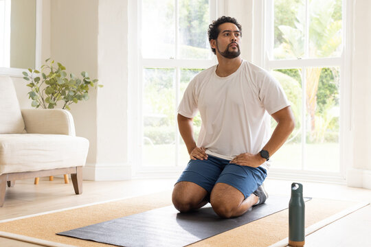 African American man in workout clothes kneeling on yoga mat in living room, holding water bottle