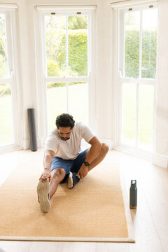 African American man in shorts stretching toward toes on mat at home with water bottle