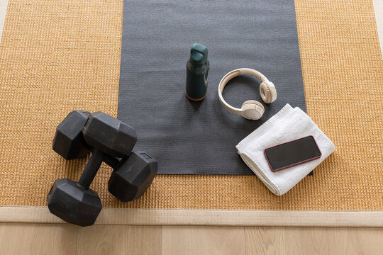 Black dumbbells are resting on gray exercise mat in home workout, with green bottle and headphones