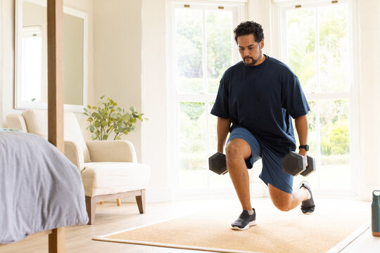 African American man lunging on rug in bedroom in shorts holding dumbbells water bottle nearby