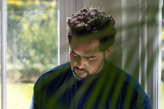 African American man looking down by window at home in blue crewneck, plant fronds shading face