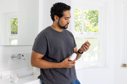 Mid-adult African American man in gray T-shirt and blue shorts, leaning at sink holding pill bottle