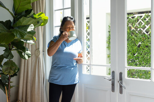 Senior Asian woman in blue T-shirt standing at home by double doors, sipping mug, holding phone