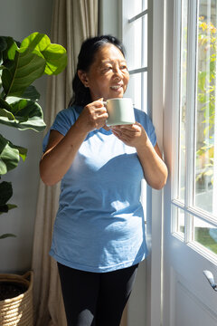 Senior Asian woman standing at glass door holding mug by plant in light-blue tee and leggings