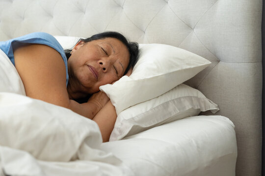 Senior Asian woman resting on left side in bedroom, wearing light blue top on stacked pillows