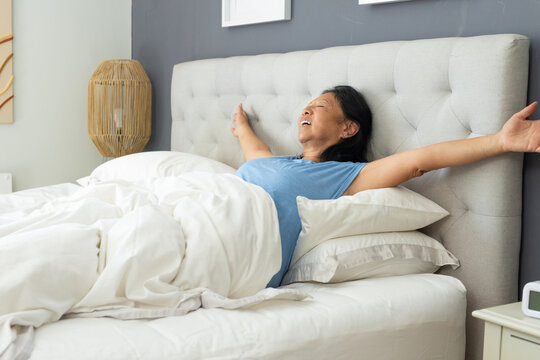 Senior Asian woman reclining and stretching arms in bed with tufted headboard wearing blue T-shirt