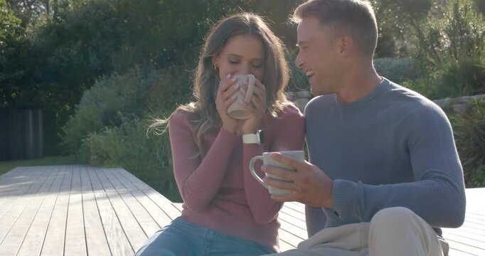 Couple sitting on wooden deck, man initiating conversation by lifting mug, woman smiling, sipping