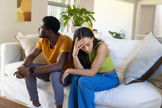 African American couple sitting on light sofa in living room, woman holding forehead, man clasping