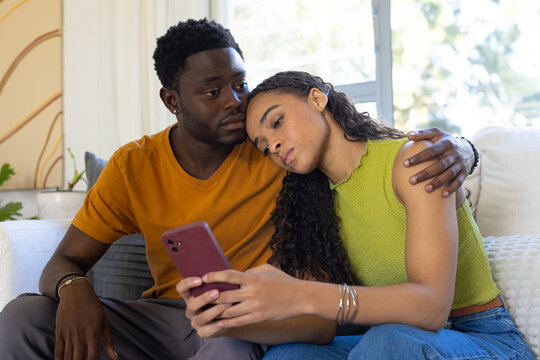 Diverse couple sitting on gray sofa at home, woman holding maroon smartphone and leaning