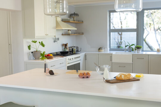 Large white kitchen island is displaying loaf, milk bottle, mixing bowl and eggs in modern kitchen