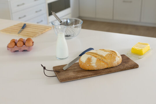 Round crusty bread is resting on wooden cutting board on white countertop, with milk, eggs, butter