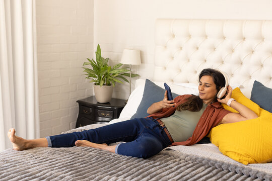 Indian woman reclining on bed wearing over-ear headphones and scrolling smartphone near nightstand