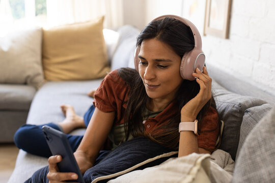 Adult Indian woman reclining on grey sofa at home, wearing pale pink headphones, holding smartphone